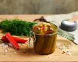 Słoik pełen pikantnych ogórków po meksykańsku, obok świeży koperek i czerwone papryczki chili.