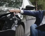 Mężczyzna w garniturze ocenia uszkodzenia czarnego samochodu po wypadku. Zastanawia się, co to AC i jak naprawić szkody.