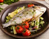Poisson entier, probablement une truite au barbecue, servi avec des tomates cerises, des quartiers de citron et des herbes fraîches sur une assiette marron.