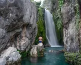 Una mujer sentada en una roca contempla la cascada de las fuentes del Algar, rodeada de vegetación y rocas.