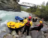 Grupa w kamizelkach ratunkowych i kaskach przygotowuje się do raftingu na rwących rzekach w Austrii. Dwie dmuchane łodzie czekają na brzegu.