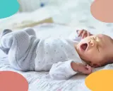 Un bebé de 1 mes bosteza, sus ojos cerrados y su boca abierta. Está acostado sobre una manta con estrellas, con las piernas cruzadas.