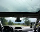 Widok przez panoramiczny dach samochodu. Krople deszczu na szybie, droga i drzewa w tle.
