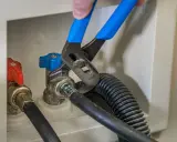 Ręka z niebieskimi szczypcami dokręca zawór do pralki. Obok czerwony zawór.