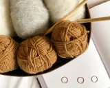 Kłębek grubej nitki w kolorze karmelowym, obok białe i szare włóczki, druty i książka o hygge.