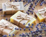 Quattro saponette fatte in casa, avvolte in carta e legate con spago, giacciono su un tavolo di legno rustico, circondate da rametti di lavanda profumata.