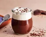 Ein Glas mit Omas Schokoladenpudding mit Sahne und Schokostreuseln. Daneben liegen ein Löffel und ein Stück Schokolade.