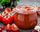 Słoik przecieru pomidorowego, świeże pomidory, czosnek i papryczki chili. Pasteryzacja przecieru pomidorowego w piekarniku to świetny sposób na jego przechowywanie.