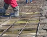 Pracownicy w gumowcach stąpają po siatce zbrojeniowej, przygotowując zbrojenie stropu do zalania betonem.