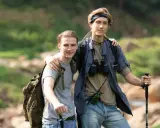 Dwóch młodych mężczyzn, gotowych na trekking, z plecakami i kijkami, w otoczeniu zieleni i skał.