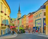 Via colorata di una città austriaca più vicina all'Italia, con edifici vivaci, una torre dell'orologio e gente che passeggia.