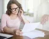 Kobieta w okularach rozmawia przez telefon, przeglądając dokumenty. Martwi się, że czeka ją brak świadectwa pracy.