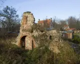 Ruiny przezmark zamku porośnięte zielenią, z widocznym fragmentem ceglanego muru z łukiem. W tle budynki i drzewa.