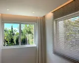 Dos tipos de apertura de ventanas: una corredera con cortinas y otra con persianas venecianas, ambas con vistas a un exuberante jardín tropical.