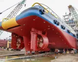 Gros navire rouge et bleu sur des supports, avec des hélices visibles. Des grues et des ouvriers sont présents. On aperçoit des anodes sur la coque, protégeant contre la corrosion, qu'elles soient anode + ou -.