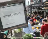 Ogłoszenie "Przyjmę do pracy" na tle ogródka restauracyjnego z ludźmi przy stolikach.