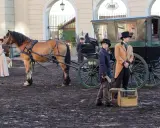 Konny zaprzęg z dorożką i postaciami w strojach z epoki na tle budynków.