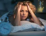 Frau mit den Händen am Kopf im Bett, offensichtlich geplagt von Kopfschmerzen direkt nach dem Aufstehen. Ein Wecker ist unscharf im Vordergrund zu sehen.