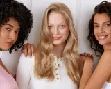 Drei Frauen stehen nebeneinander. Die Frau in der Mitte hat orange blonde haare und lächelt.