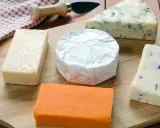 Verschiedene Käsesorten auf einem Holzbrett. Warum ist Cheddar orange? Die natürliche Farbe kommt von Beta-Carotin, das Kühe über Gras aufnehmen.