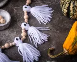 Niedliche Geister-Girlande zum Halloween basteln, mit Holzperlen und weißen Fransen. Daneben liegen bunte Kürbisse.