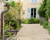 Un chemin de gravier serpente à travers un jardin anglais romantique, bordé de roses roses et de verdure luxuriante. Une maison en pierre avec des volets bleus se profile à l'arrière.