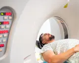 Pacjent leży w aparacie do tomografii komputerowej, przygotowując się do badania. Obok widoczny panel sterowania z cyfrowymi wskaźnikami.