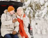 Ett äldre par sitter i bakluckan på en bil i snöig skog, njuter av livet med varma drycker.