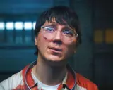 Paul Dano med glasögon och blodfläckar på sin randiga skjorta, i en mörk miljö med ljus bakom.