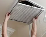 Man hands removing a pleated air filter from a ceiling vent. Essential for limpiar filtros aire acondicionado.