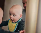 Ein Baby mit gelbem Lätzchen schaut überrascht in einen Spiegel. Vielleicht fragt es sich, ab wann Kinder "Ich" sagen.