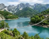 Un ponte sospeso attraversa il lago di Vagli, con montagne imponenti e un borgo pittoresco sullo sfondo.