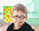 Niño con gafas y parche en el ojo, tratando su ojo vago. Detrás, una pantalla con un patrón de colores.