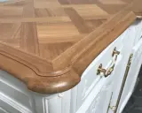 Un meuble ancien relooké en moderne, avec un plateau en bois clair à motif géométrique et des poignées dorées sur un corps blanc.