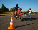 Persona con casco y equipo de protección conduce una moto naranja en un circuito de prácticas. Un instructor observa.