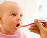 Niemowlę z niebieskimi oczami otwiera buzię na łyżeczkę z jedzeniem. To jego pierwszy posiłek niemowlaka po 4 miesiącu.