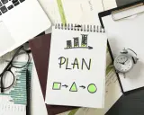Na biurku leżą dokumenty, laptop, okulary i budzik. Na notatniku widnieje napis "PLAN" i schemat, który symbolizuje indywidualny plan rozwoju.