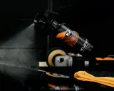 Ręka w czarnej rękawiczce rozpyla płyn K2 Klinet, środek do inspekcji lakieru z alkoholem IPA. W tle polerka i pomarańczowa mikrofibra.