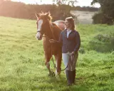 Frau geht mit braunem Pferd spazieren, das Halfter trägt. Sie genießen einen sonnigen Tag auf der Wiese.