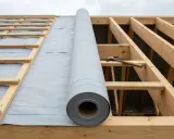 Rouleau de membrane HPV sur charpente de toit en bois. Un marteau est posé sur une poutre.