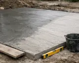 Świeżo wylany beton, widoczna różnica w wilgotności i fakturze. Narzędzia budowlane obok sugerują początek prac i testowanie wytrzymałości betonu po 1 dniu.