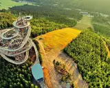 Drewniana ścieżka w koronach drzew w Czechach, przypominająca spiralny sky bridge, otoczona lasem i złotym polem.