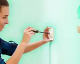 Une femme souriante utilise un tournevis pour installer une prise électrique. Un personnage de dessin animé en casque vert fait un clin d'œil et un signe "OK".
