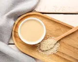 Eine Schale cremiger Tahin neben Sesamsamen auf einem Holzlöffel. So sieht die Antwort auf die Frage "was ist Tahin" aus.