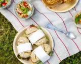 Idée pique-nique : petits pains garnis, salades en bocaux et muffins sur une nappe rayée, posée sur l'herbe.