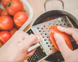 Ręce trą pomidora na tarce, przygotowując go do sosu. Obok stoi miska pełna świeżych pomidorów.