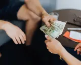 Dwie osoby przekazują sobie plik banknotów. Scena sugeruje nielegalną transakcję, być może korupcję.