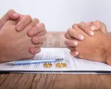 Dwie pary splecione dłonie nad dokumentem, na którym leżą obrączki i długopis. Może to symbolizować trudne decyzje, jak kredyt przed ślubem.