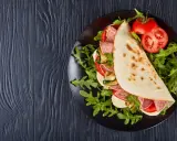 Piadina ripiena con salame, mozzarella, zucchine grigliate e pomodoro, servita su un piatto nero con rucola. Un assaggio della città di Rimini in un ristorante.