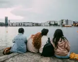 Three young people sit on rocks by the water, looking at a city skyline.